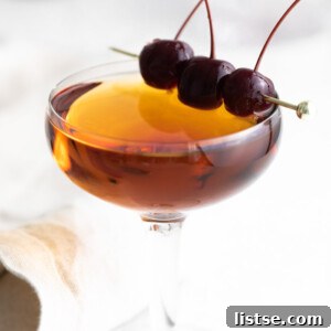 Gotham's Ruby 5 Manhattan cocktail (brownish orange liquid) in a coup cocktail glass and garnished with 3 dark maraschino cherries on a gold pick