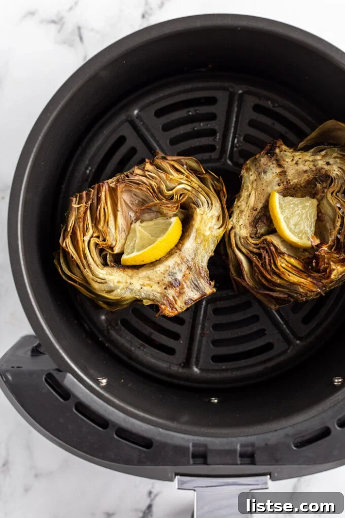 Close-up of cooked artichoke halves arranged in the basket of an air fryer, with lemon wedges for garnish.