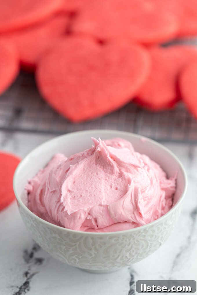 Delicious pink sugar cookie frosting in a bowl surrounded by festive red cookies, perfect for holiday decorating.