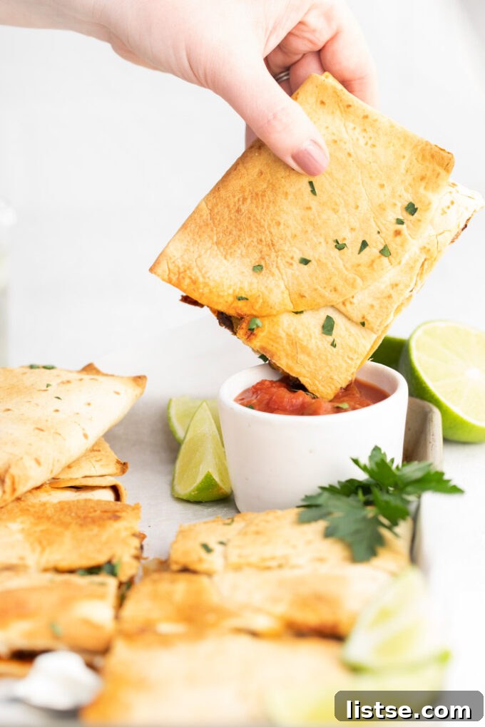 A square of golden sheet pan quesadilla being dipped into a small bowl of fresh salsa, with more quesadillas in the background.