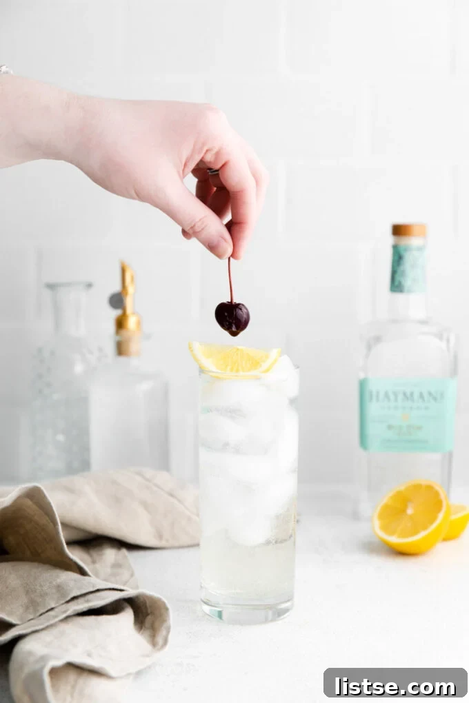 tom collins cocktail in a tall glass with ice and garnished with a lemon wedge and dark cherry. Liquor bottles in the background. hand placing the cherry on top of the drink