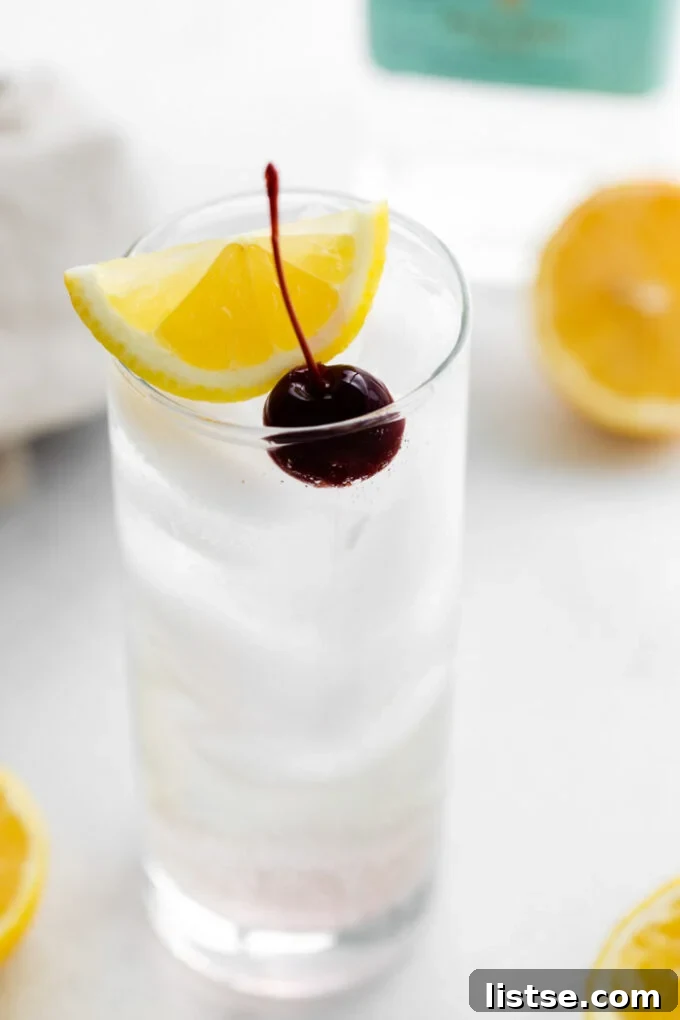 side shot of a tom collins (clear liquid) in a tall glass filled with ice and garnished with lemon wedge and dark cherry