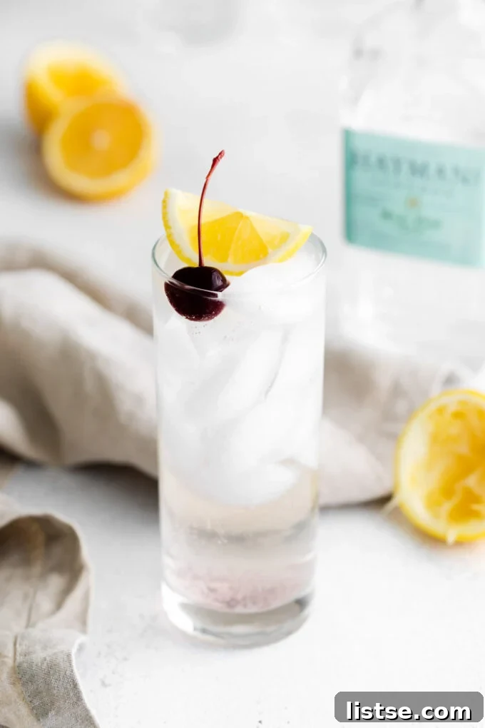 tom collins cocktail in a tall glass with ice and garnished with a lemon wedge and dark cherry. Liquor bottles in the background