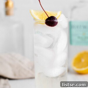 tom collins cocktail in a tall glass with ice and garnished with a lemon wedge and dark cherry. Liquor bottles in the background