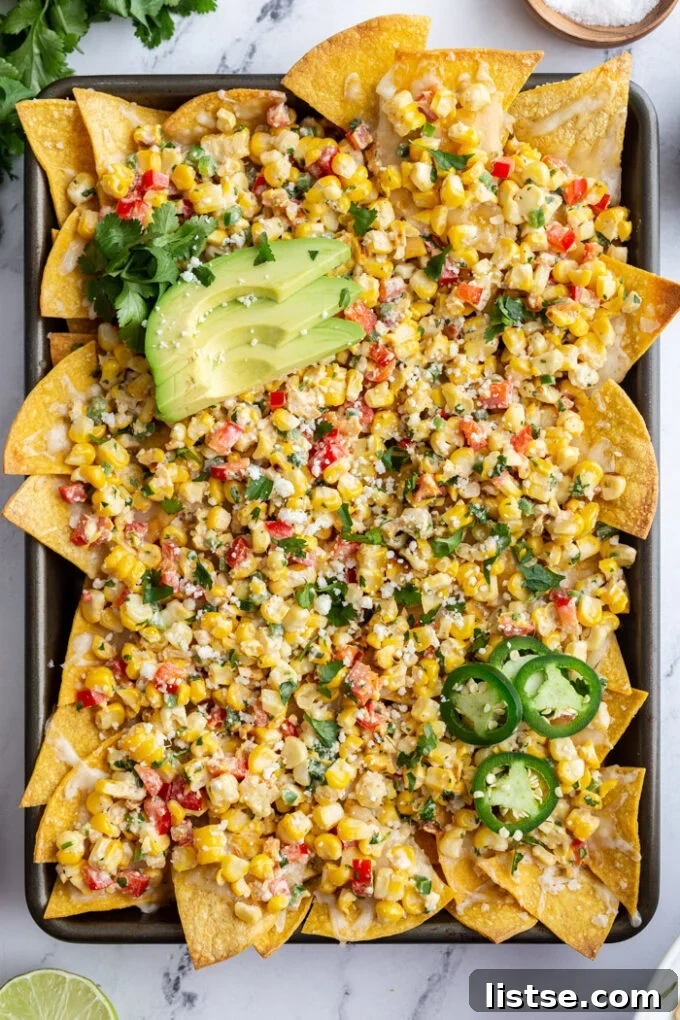 Elote Nachos 2 A baking tray filled with golden tortilla chips covered in melted cheese and a vibrant Mexican street corn salad, garnished with fresh cilantro and lime wedges.