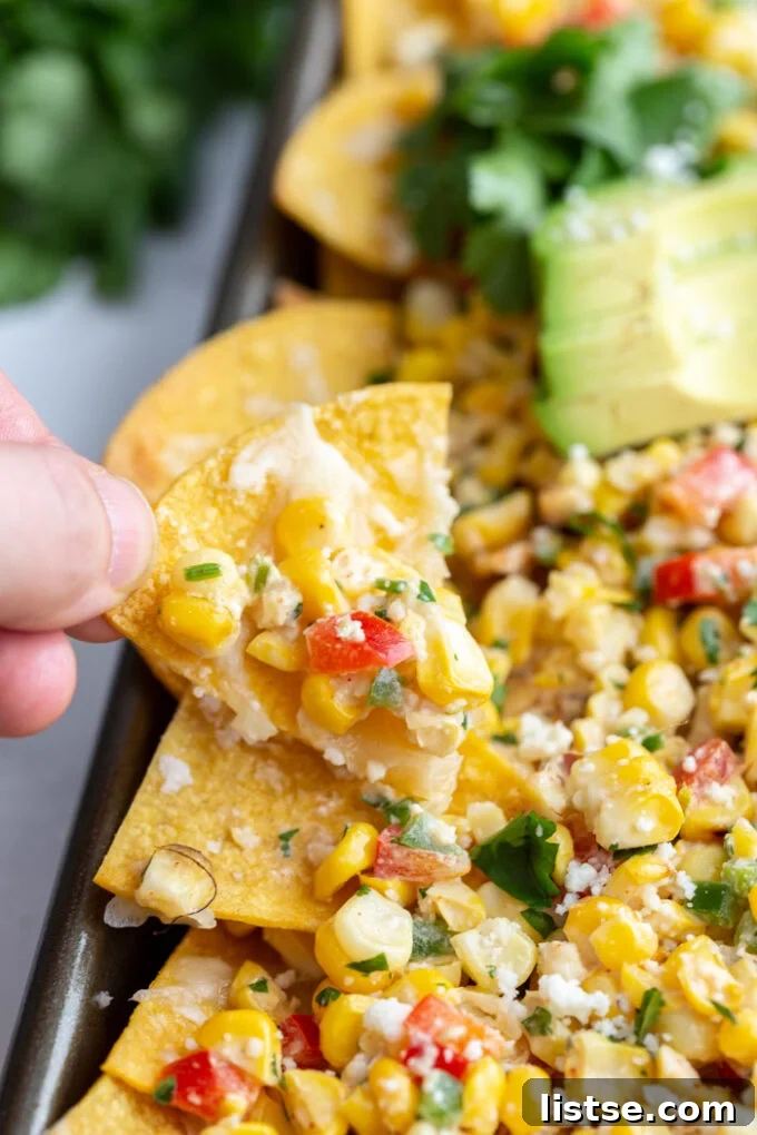 Elote Nachos 3 A hand holding a single tortilla chip loaded with creamy street corn salad, with a full tray of nachos blurred in the background.
