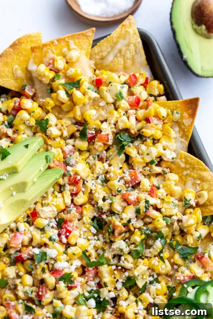 Elote Nachos 7 A tempting side angle view of a baking tray laden with colorful Street Corn Nachos, with a fresh avocado half in the soft-focus background, suggesting additional garnishes.