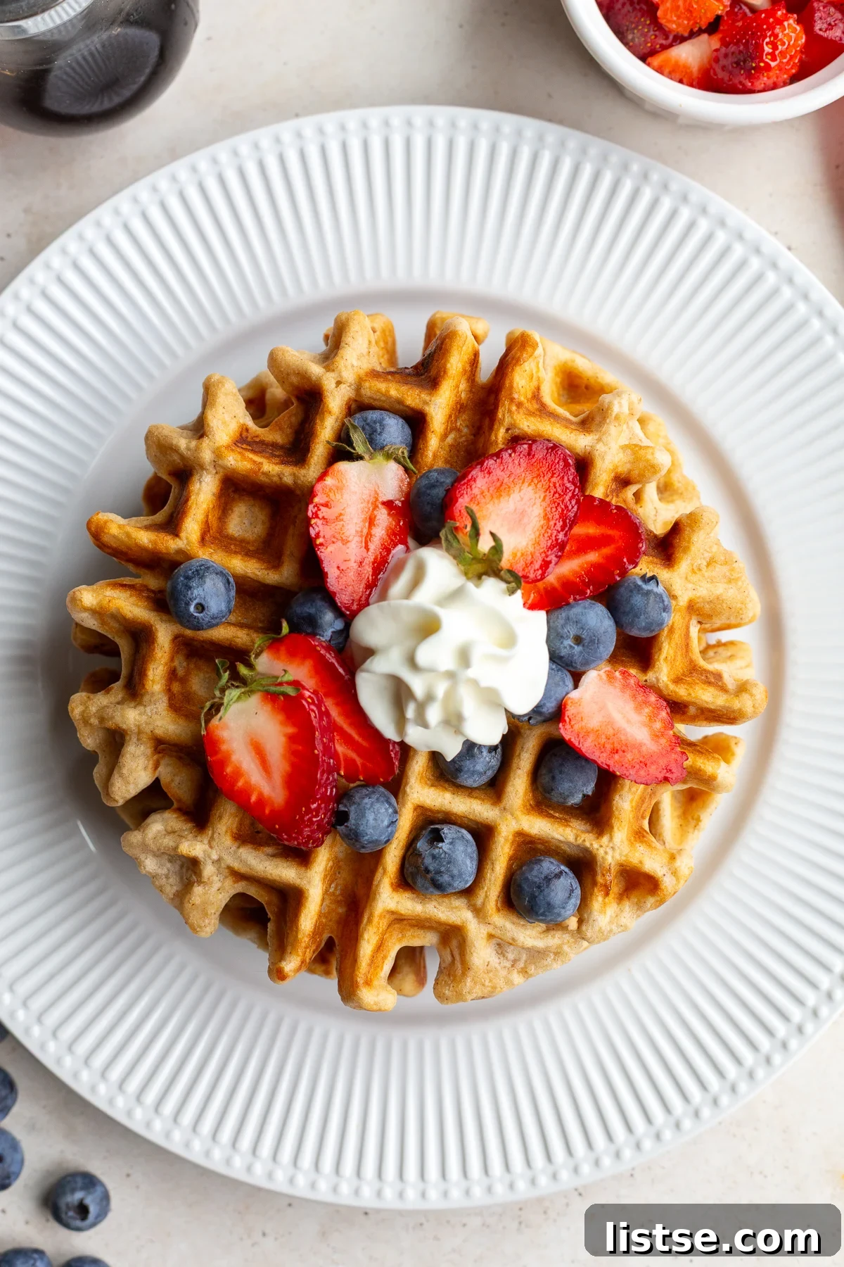 A golden vegan waffle on a pristine white plate, beautifully garnished with vibrant fresh berries and a dollop of non-dairy whipped cream, ready to be enjoyed.