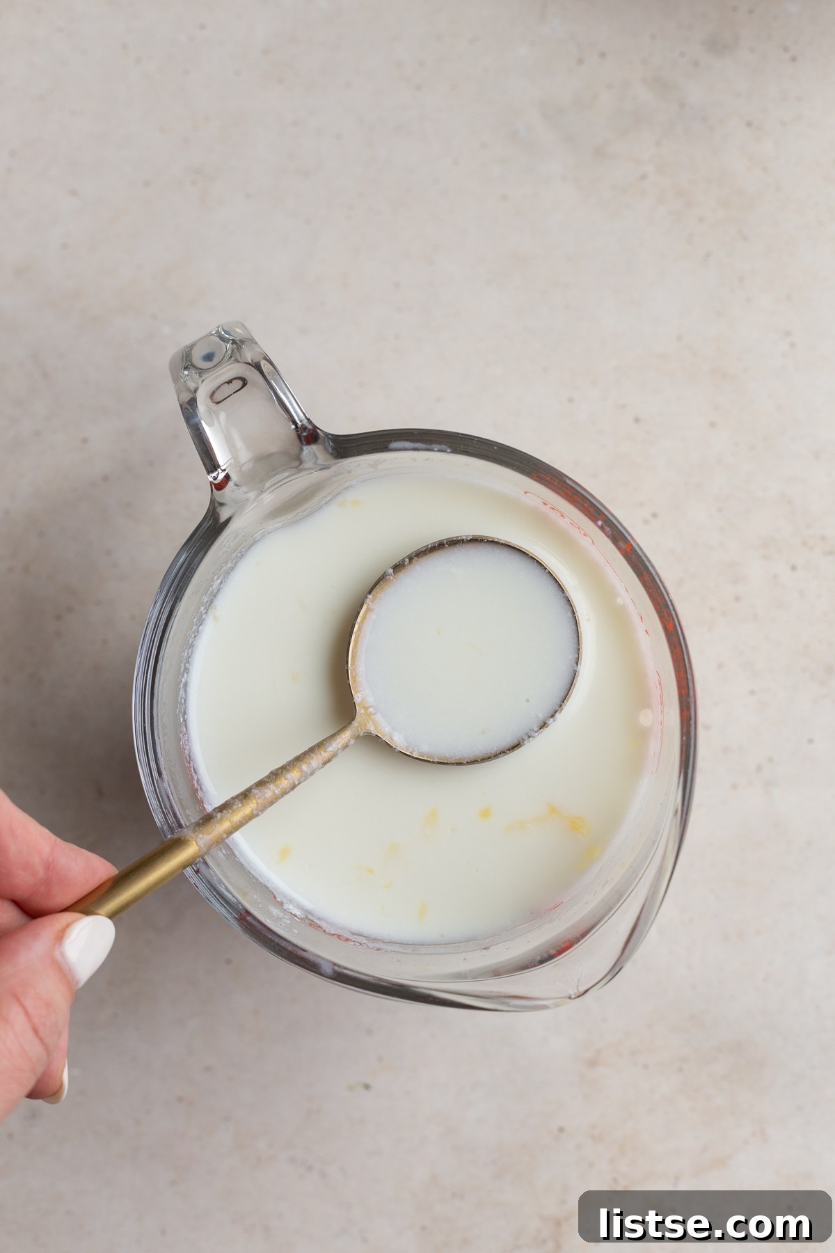 Using a spoon to gently stir dairy-free milk and fresh lemon juice in a spouted measuring glass, forming vegan buttermilk.