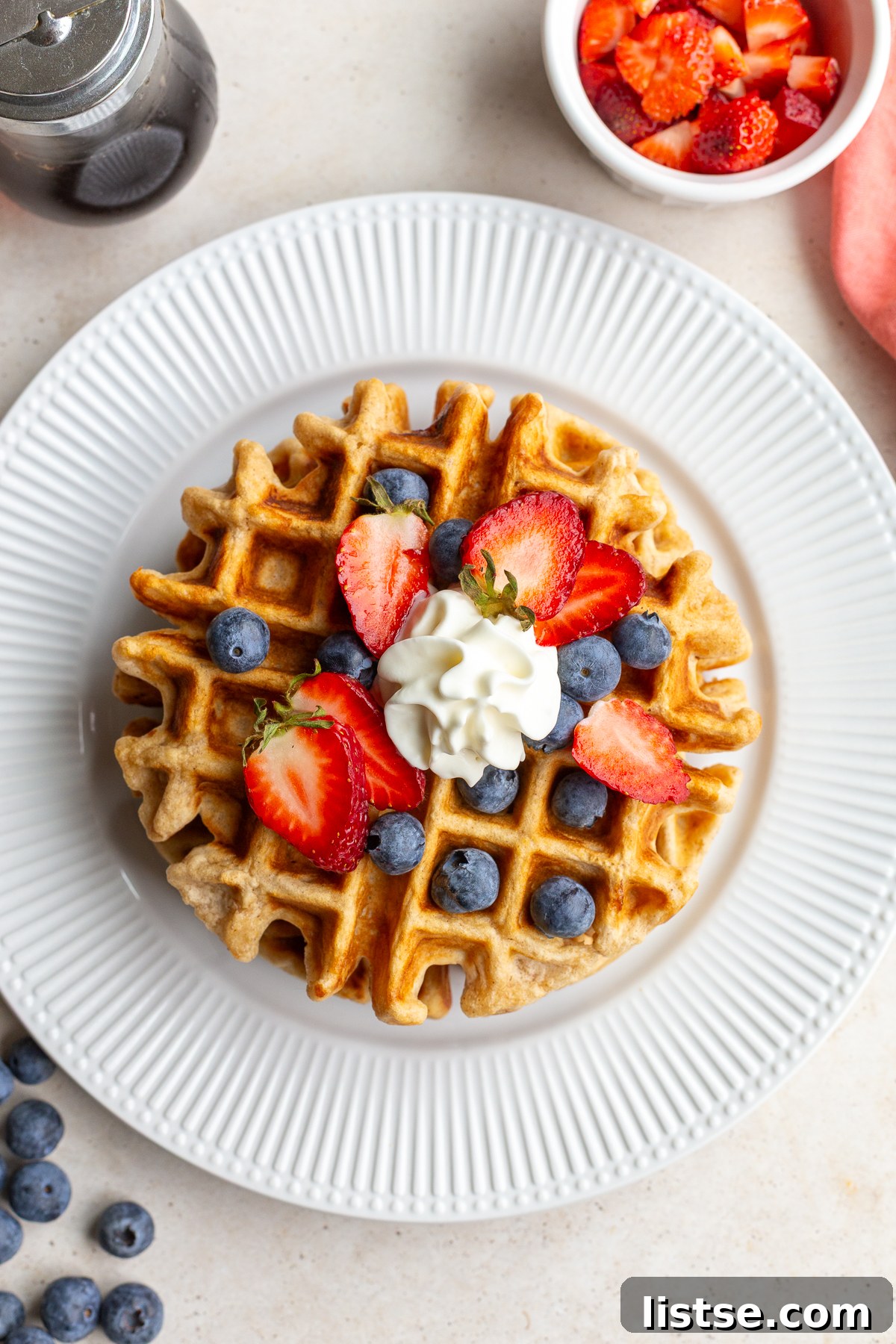 A stack of golden vegan waffles, generously topped with an assortment of fresh strawberries, blueberries, and a dollop of fluffy whipped cream, ready to serve.