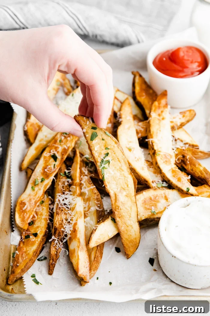 Crispy potato wedges arranged on a parchment-lined baking sheet, sprinkled with cheese and herbs, alongside ketchup and ranch, with a hand holding one wedge