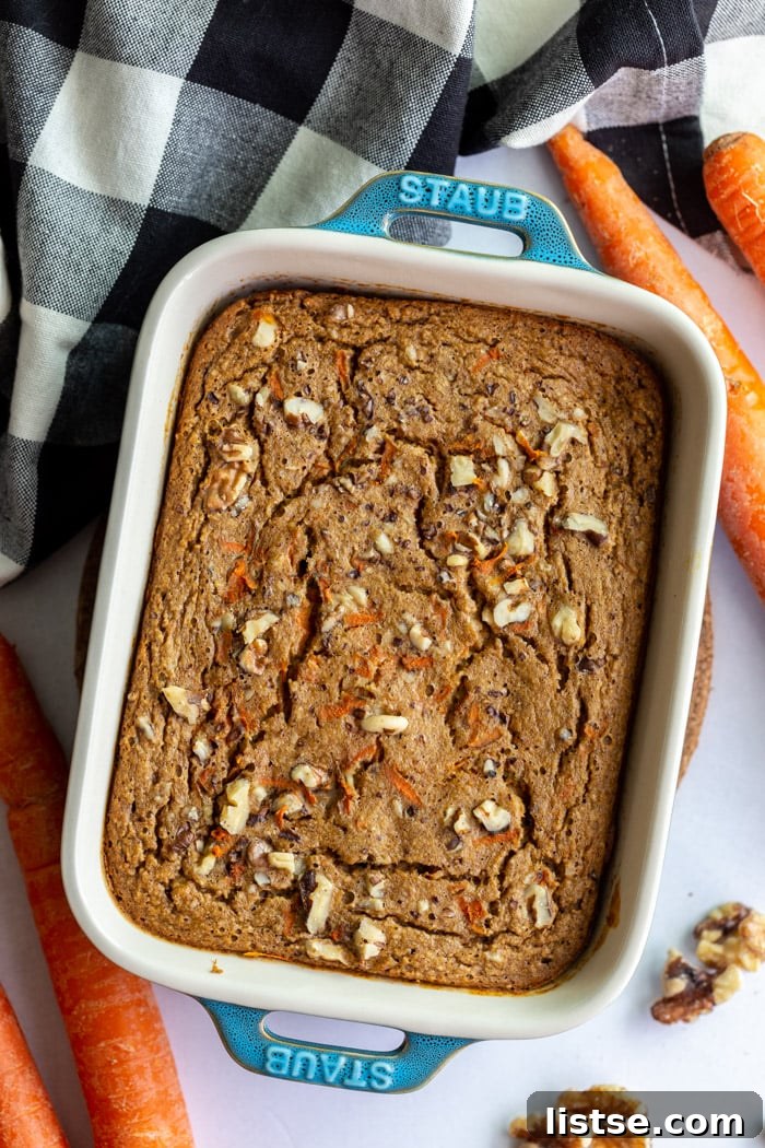 Green casserole dish filled with baked carrot cake oatmeal, with fresh carrots and a towel on the side.