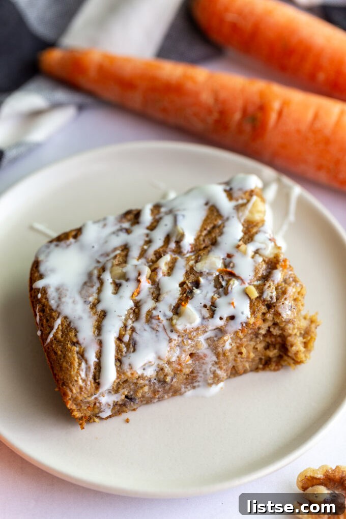 A slice of carrot cake baked oats on a plate with cream cheese frosting drizzled on top, with carrots in the background.