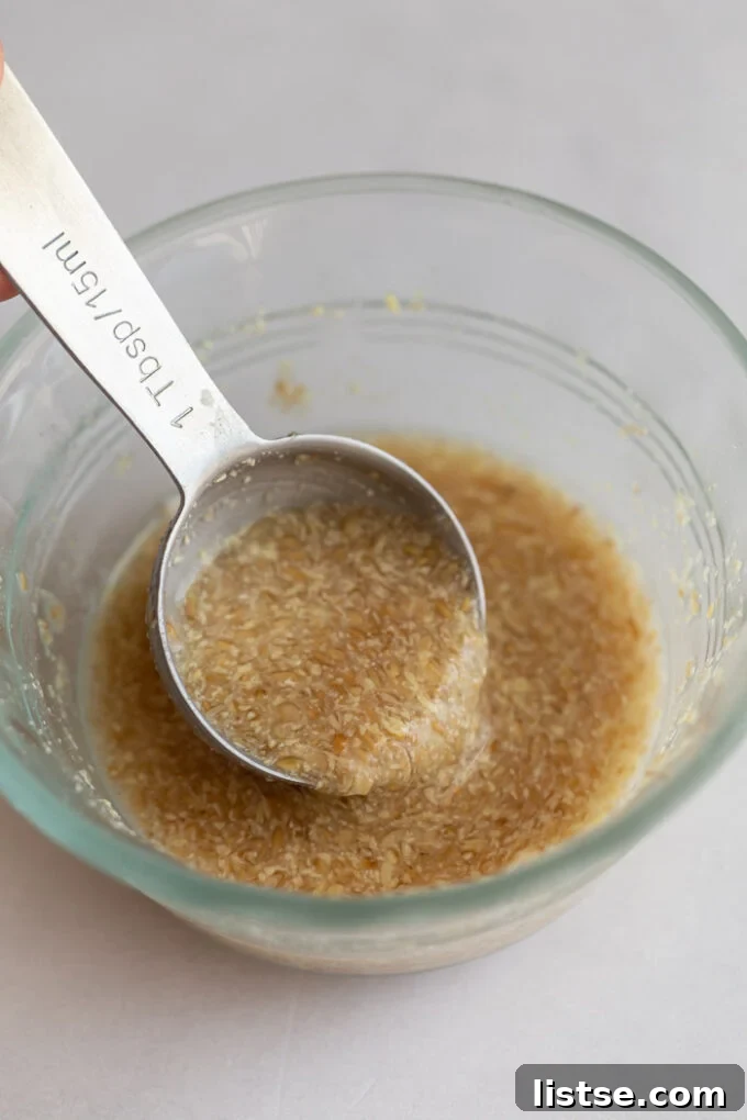 A glass bowl holds a smooth, gel-like flax egg, with a metal spoon gently scooping a portion, illustrating its readiness for baking.