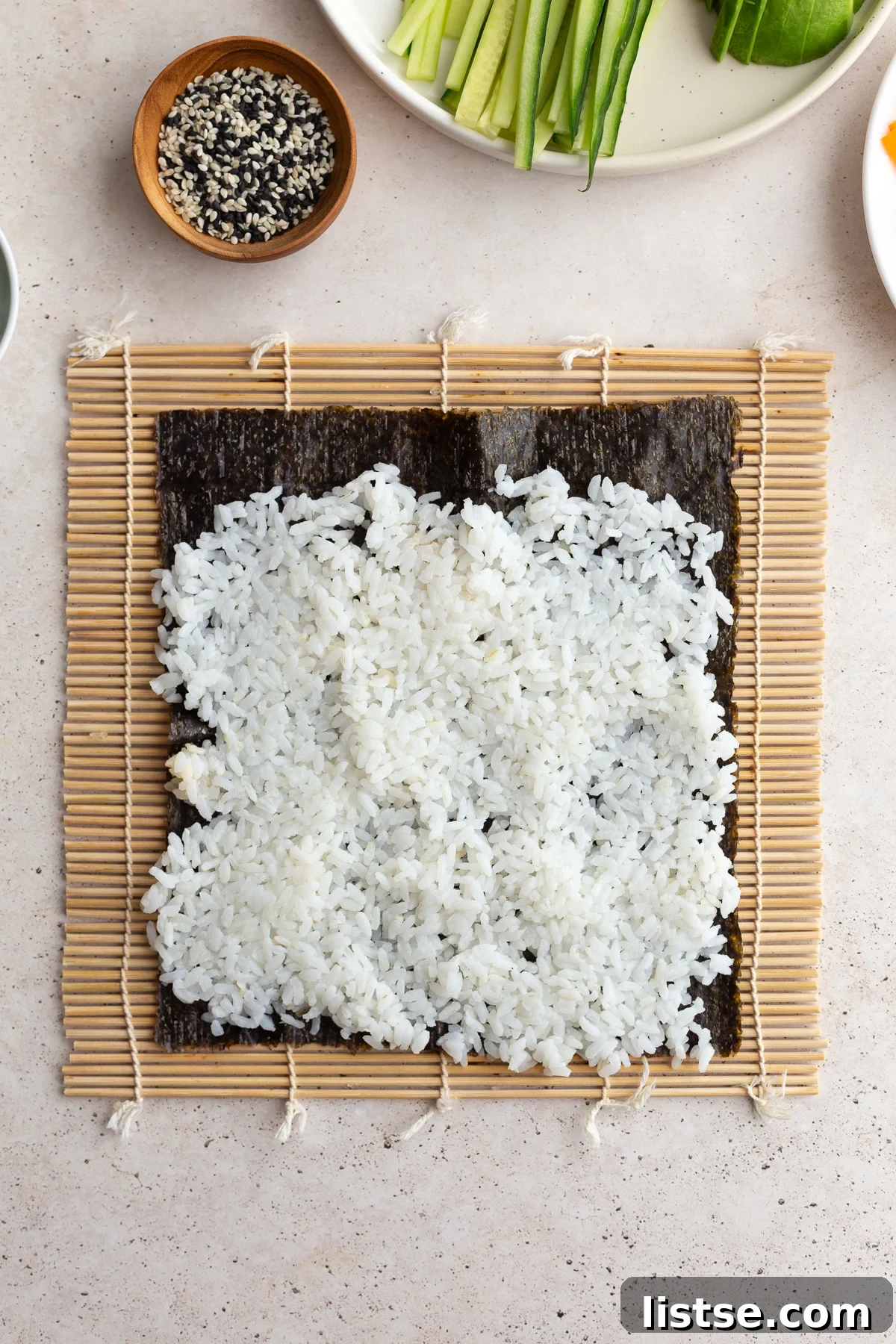 A sheet of nori placed on a sushi rolling mat, being topped with a layer of prepared sushi rice.