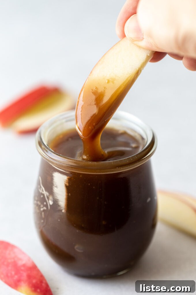 A hand dipping a fresh apple slice into a small glass jar filled with luscious vegan caramel sauce, showing its inviting texture.