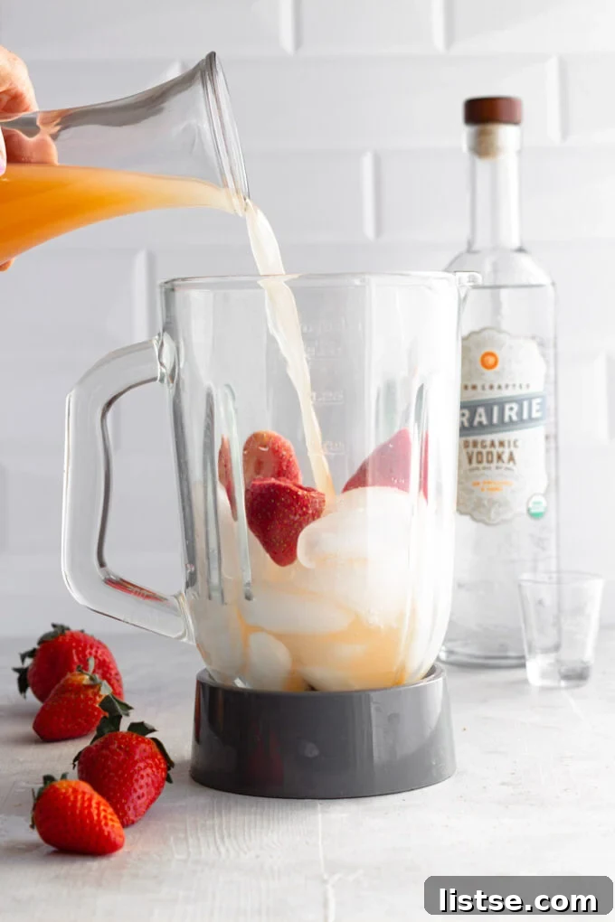 Blender filled with ice and fresh strawberries, with grapefruit juice being poured in, illustrating the cocktail preparation.