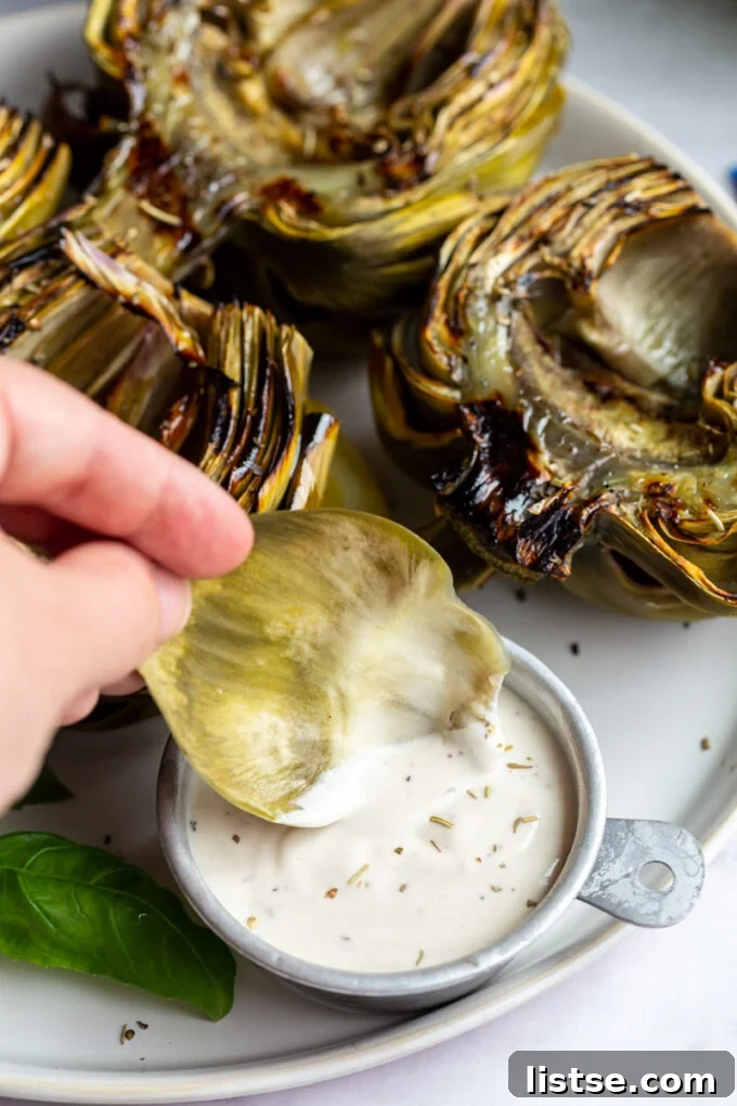 Fire-Kissed Artichokes 4 A hand holding a grilled artichoke leaf, dipping it into a creamy sauce, with a plate of grilled artichokes in the background.