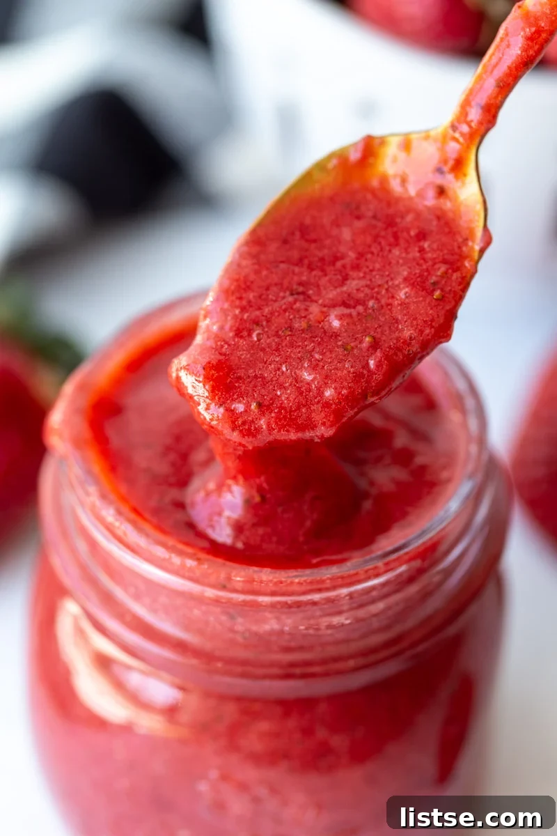 A spoon scooping fresh strawberry puree from a small glass jar, showcasing its smooth texture and vibrant red color.