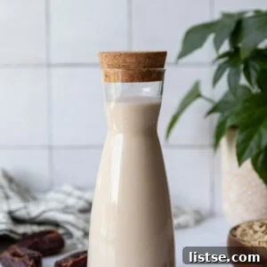 Tall clear container of oat milk with fresh dates and oats around it and a fresh plant in the corner