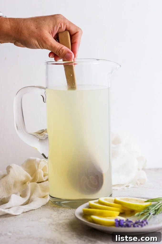 A wooden spoon stirring lavender lemonade in a glass pitcher, ensuring all ingredients are well combined.