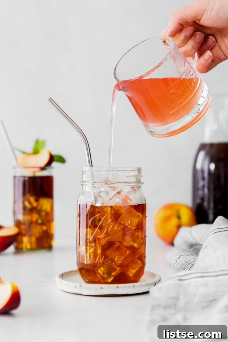 Close-up shot of a hand pouring vibrant homemade peach simple syrup into a glass filled with ice and amber-colored brewed tea, creating a beautiful layered effect.