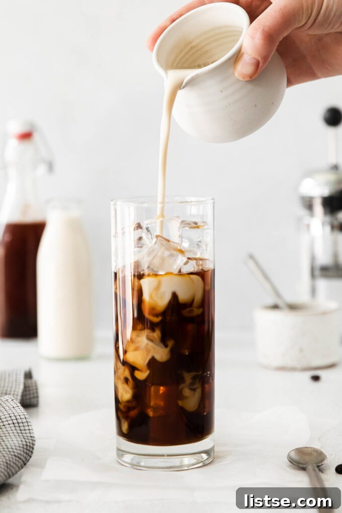 A tall glass of iced coffee, with creamy oat milk being poured in, demonstrating the layering effect of a delicious latte.