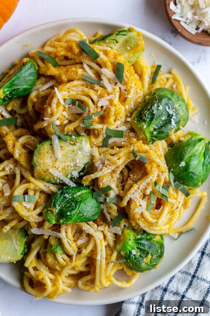 A large plate of spaghetti generously coated in a rich pumpkin sauce, crowned with perfectly halved and tender Brussels sprouts. Small decorative pumpkins are artfully placed in the corner of the shot, hinting at the autumnal theme. A bag of Ocean Mist Farms Quick Cook Sprouts peeks into the frame, emphasizing the ease of preparation.
