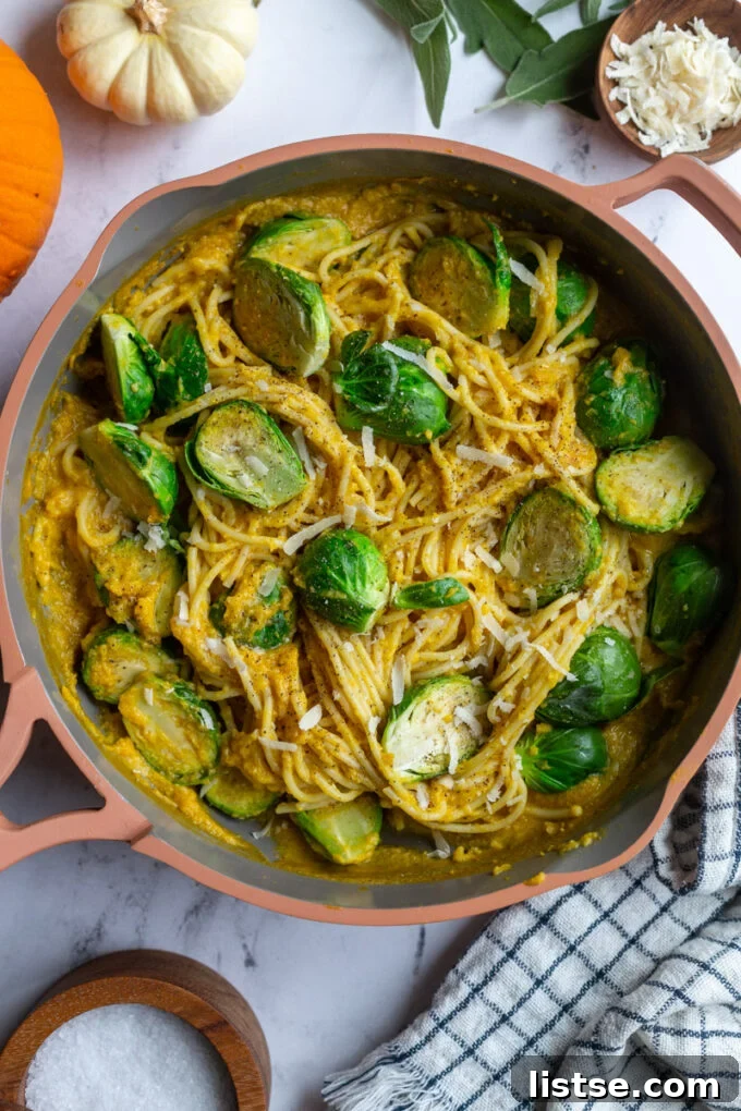 A close-up shot of creamy pumpkin pasta spaghetti simmering in a large skillet, topped with tender Brussels sprouts. Small decorative pumpkins are visible in the background, reinforcing the seasonal theme.