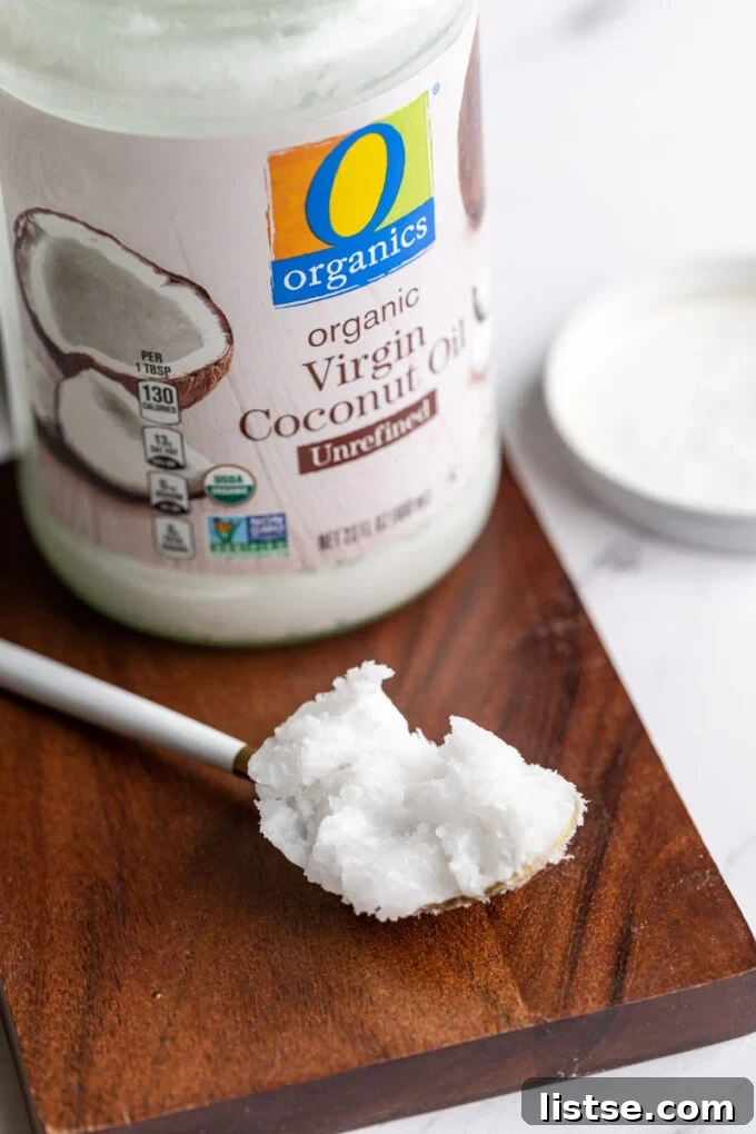 A jar of solid coconut oil on a wooden board, with a spoonful of melted coconut oil resting beside it, indicating its versatile states.