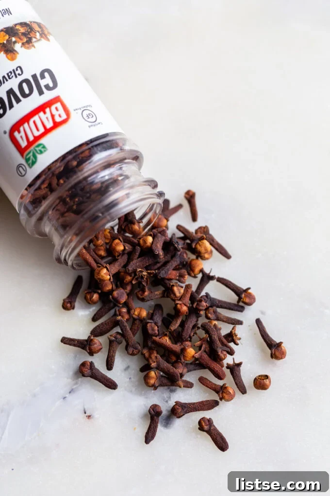 A glass jar filled with whole cloves spilled onto a wooden surface, highlighting their distinctive spike and bulbous top.