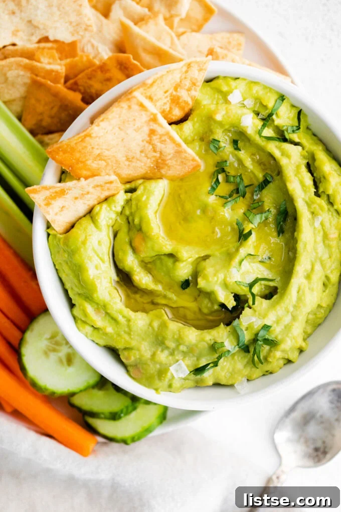 Inspired Chickpea Meals 12 A white bowl brimming with vibrant green avocado hummus, artfully surrounded by an assortment of chips and fresh vegetables, with a few chips dipped in.