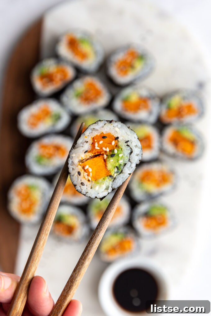 A hand holding a perfectly rolled sweet potato sushi piece with chopsticks, showcasing the vibrant filling and crisp exterior.