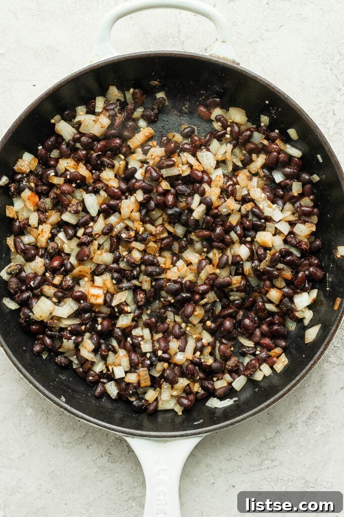 Cooking black beans with diced onion and spices in a large skillet, forming a savory base for vegetarian nachos.
