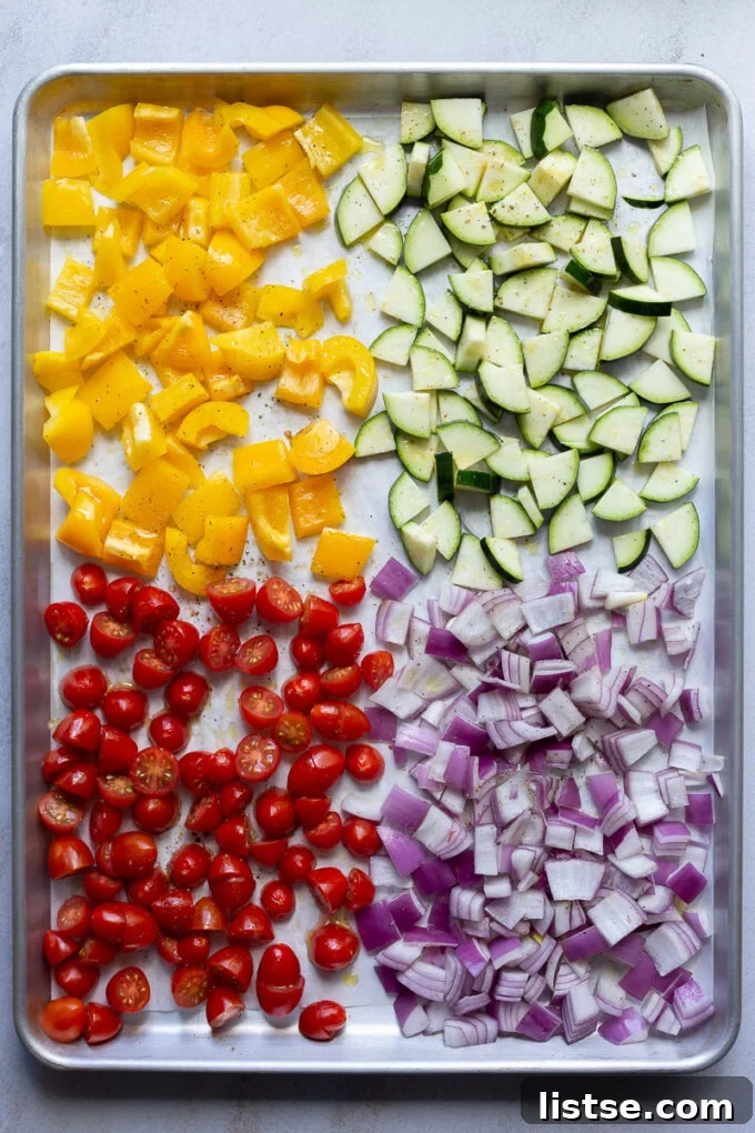Raw bell pepper, zucchini, red onion, and cherry tomatoes on a baking pan, ready for roasting.