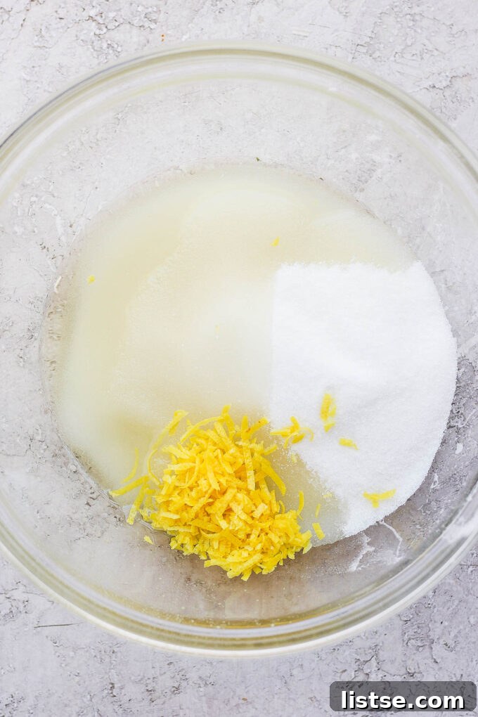 A close-up shot of lemon zest being mixed with sugar and oil in a glass bowl, illustrating the initial wet ingredients step.