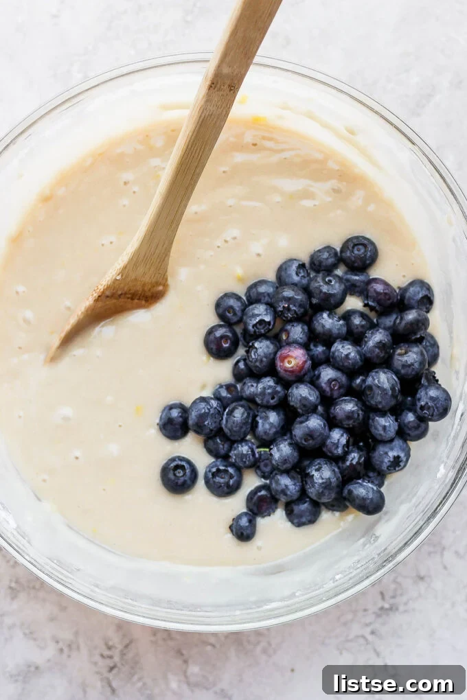 Muffin batter with a generous amount of blueberries being gently folded in with a spatula, highlighting the careful mixing process.