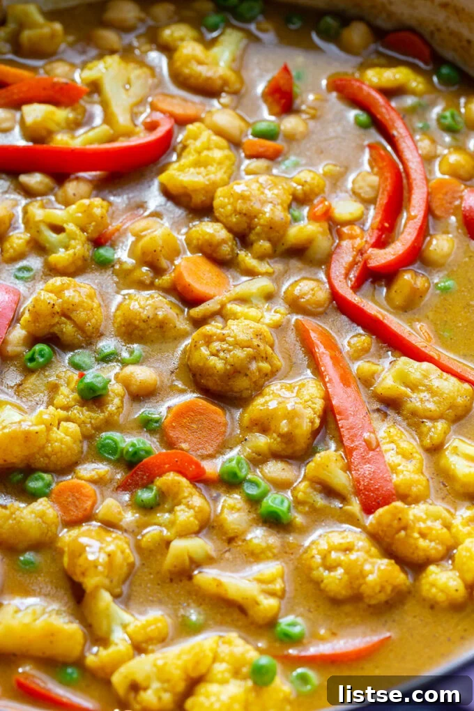Close-up of cauliflower and other vegetables simmering in a rich, orange-red curry sauce in a large pan.