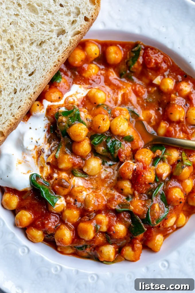 A bowl of rich, red braised chickpeas, garnished with fresh herbs and served alongside a slice of crusty bread and a spoonful of creamy yogurt.