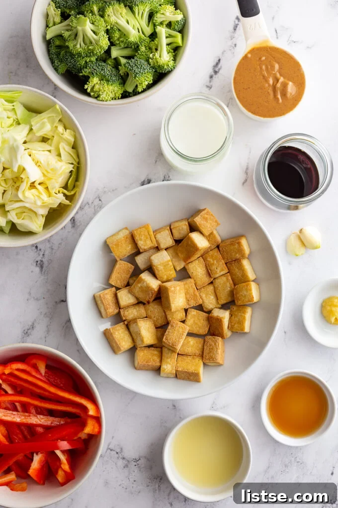 A collection of ingredients for Thai Peanut Sauce Tofu, neatly arranged in small bowls: pressed tofu, olive oil, peanut butter, soy sauce, garlic, milk, maple syrup, lime juice, ginger, bell pepper, broccoli, and cabbage.