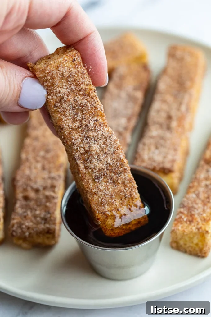 A woman's hand gracefully dipping a French toast stick into a small bowl of golden maple syrup.