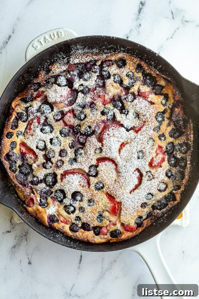 A whole Berry Dutch Baby pancake in a white skillet, dusted with powdered sugar, ready to be portioned and served for a delightful family breakfast.