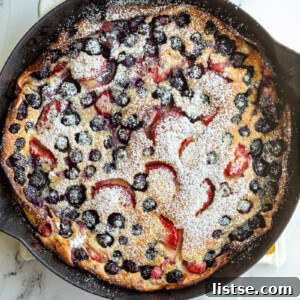 A vibrant Berry Dutch Baby pancake in a white skillet, garnished with fresh blueberries and strawberries, and lightly dusted with powdered sugar.