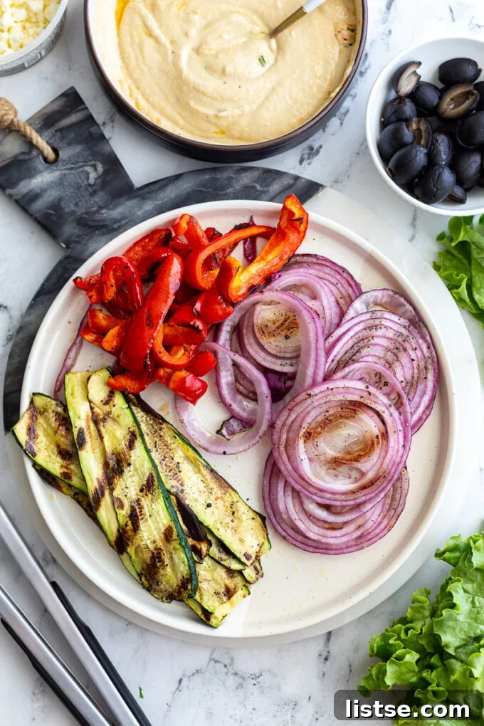 A vibrant display of fresh and grilled ingredients for veggie wraps, including grilled zucchini, bell pepper, and onion, alongside a bowl of hummus, black olives, and fresh lettuce.
