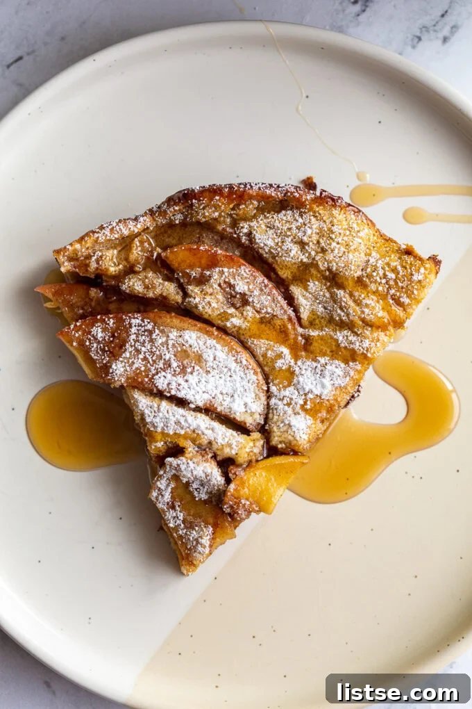 Golden Apple Oven Puff 3 A single slice of Apple Dutch Baby on a white plate, elegantly dusted with powdered sugar and drizzled with maple syrup.
