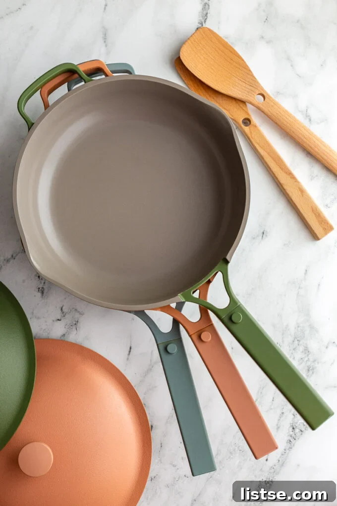 Three Our Place Always Pans in blue, pink, and green stacked on top of each other with wooden spoons and lids in the corner