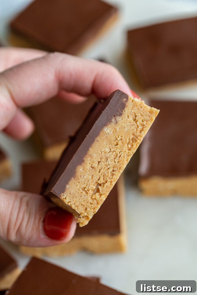 A hand holds up a single perfect chocolate peanut butter bar with a clear top layer, with more delicious bars visible in the background, ready to be enjoyed.