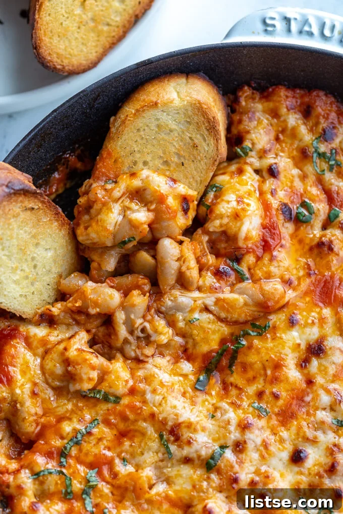Close-up of Cheesy White Bean Tomato Bake in a white cast iron skillet, with a serving scooped onto toasted bread, garnished with fresh basil.