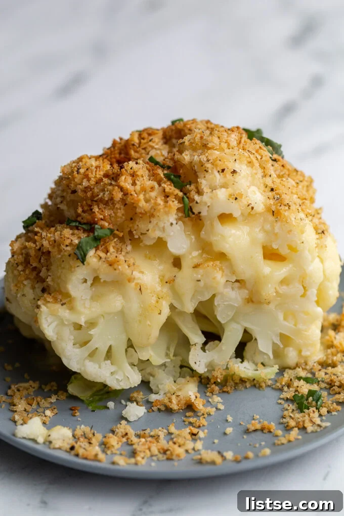A whole head of cheesy stuffed cauliflower, cut in half, revealing melted white cheddar cheese oozing out, garnished with toasted breadcrumbs and fresh basil ribbons, resting in a blue baking dish.