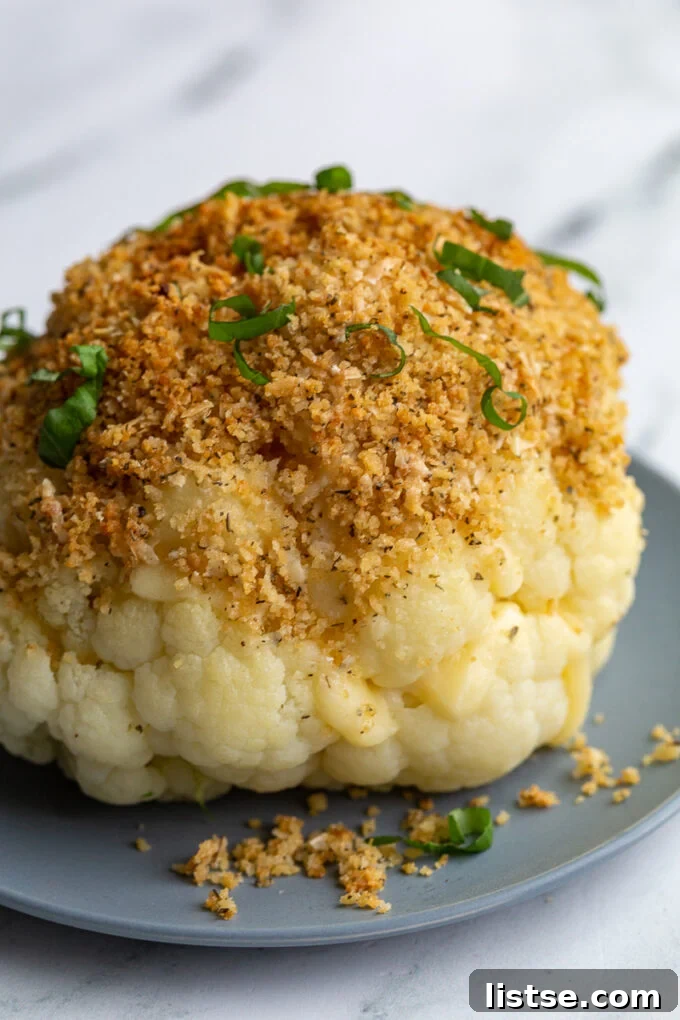 A perfectly roasted whole head of cheesy stuffed cauliflower in a blue dish, with melted white cheese peeking from the florets, crowned with a golden toasted breadcrumb mixture and delicate basil leaves.